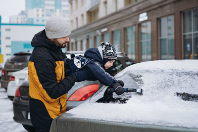 Boy wearing scandinavian knit hat helping to brush snow from a car