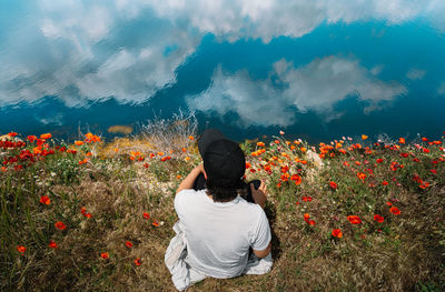 Man sitting in poppy field by the lake with clouds reflection