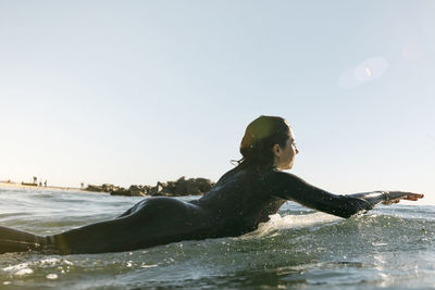 Side view of female surfer lying on surfboard in sea against clear sky