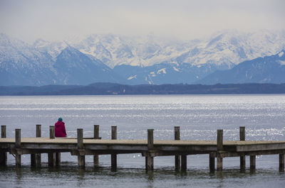 Person sitting on pier over sea against snowcapped mountains