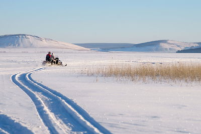The snowmobile with fishermen is riding along the winter lake against the backdrop of the village.
