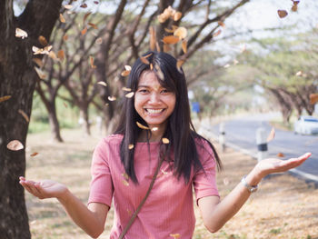 Portrait of smiling young woman throwing leaves and twigs while standing against trees