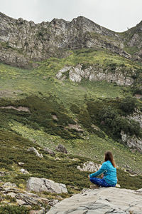 Rear view of meditating woman in blue sitting on stone in mountain, meditation in nature mindfulness