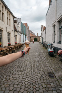 Cropped image of hand holding ice cream cone on street amidst buildings