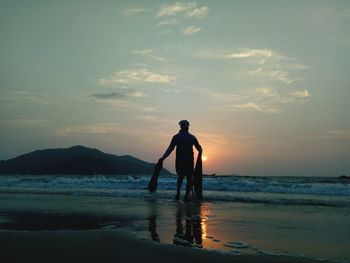 Silhouette fisherman on shore at beach against sky during sunset