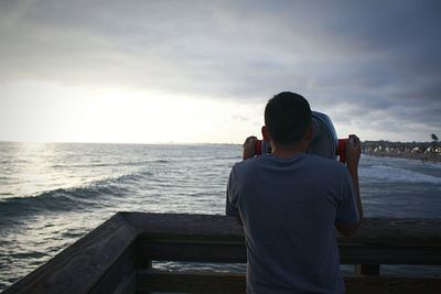 Rear view of man looking through binoculars by sea against cloudy sky