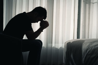 Side view of silhouette man sitting against window at home