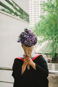 Person covering face with bouquet while wearing graduation gown against buildings in city