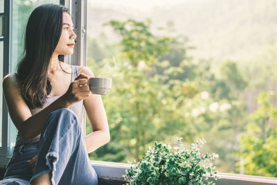 Young woman drinking coffee while sitting on window