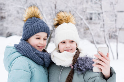 Two smiling girls in warm coats and hats are photographed on a smartphone in a snowy winter park. 