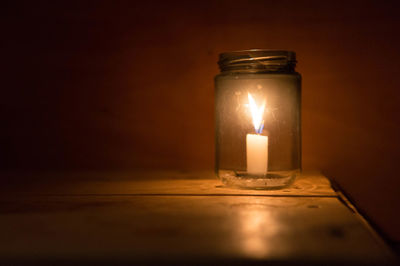 Close-up of illuminated candle in glass jar on table against wall