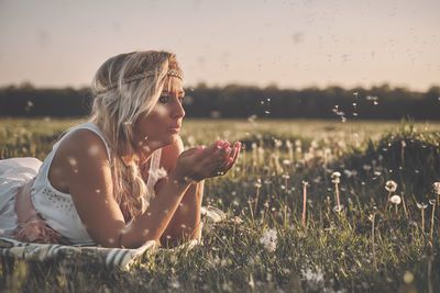 Young woman blowing dandelions while lying on field during sunset