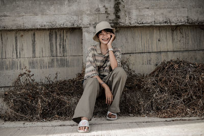Full length portrait of smiling woman sitting against wall outdoors