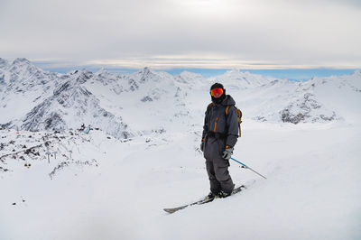 Young man stands on a rock at the top of a mountain with a backpack and skis, watching a long alpine