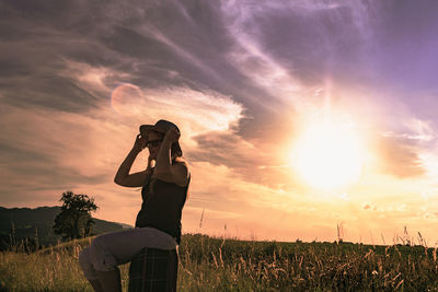 Side view of woman sitting on luggage at grassy field during sunset