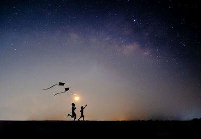 Silhouette people flying kite on field against sky at dusk