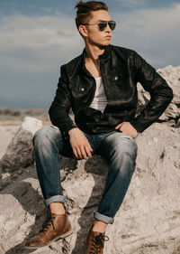 Full length of fashionable young man looking away while sitting on rock