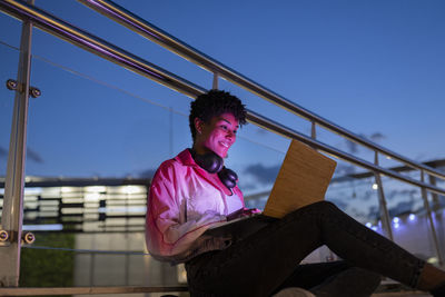 Young woman using laptop while sitting by glass railing during night