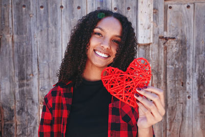 Portrait of cheerful woman holding red heart shape against wooden wall