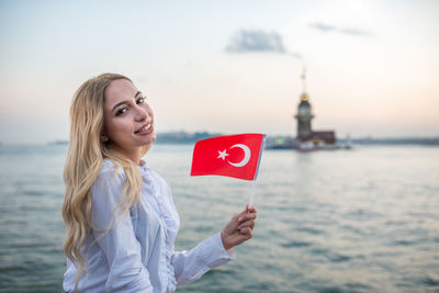 Portrait of young woman with blond hair holding turkish flag against sea