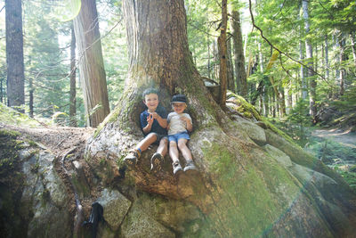 Two young explorers sit below old growth tree in natural forest.