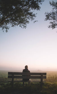 Rear view of man sitting on bench against landscape during foggy weather