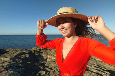 Portrait of a caucasian young woman in a red dress and straw hat on the seashore. holds a hat with 