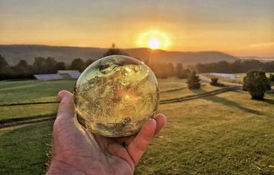 Close-up of hand holding crystal ball on field against sky during sunset