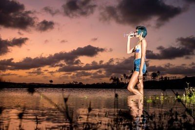 Side view of young woman drinking wine while standing in lake during sunset