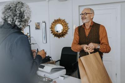 Smiling senior male entrepreneur giving paper bag to female customer while shopping in store