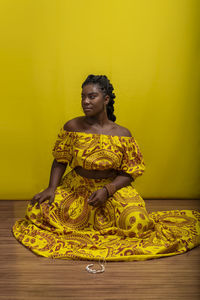 Young woman wearing colorful long clothes sitting over yellow background. natural expression.