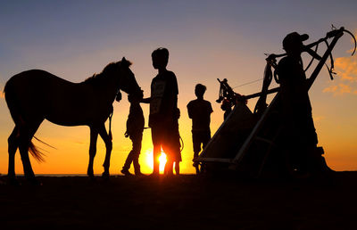 Silhouette boys with horse at cart at beach against sky during sunset