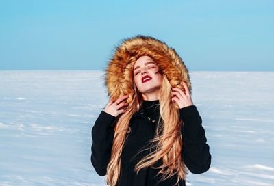Beautiful young woman wearing fur coat standing in snow against clear sky