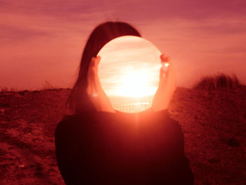 Woman holding mirror against face while standing outdoors during sunset