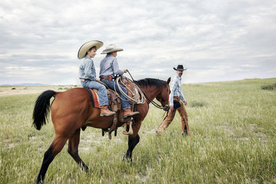 Father looking at sons riding horse on field against cloudy sky