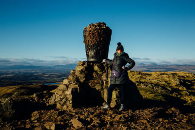 Full length portrait of woman standing by monument against blue sky at dumyat