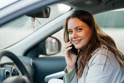 Beautiful girl with long hair in a grey trench coat using smartphone call gets into the car