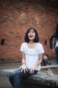 Young woman with mouth open throwing food while sitting against brick wall