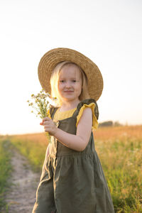 A little blonde girl in a straw hat walks in a field with a bouquet of daisies. 