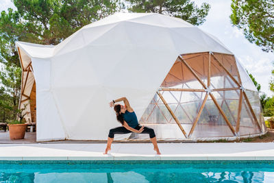 Unrecognizable fit female in sportswear standing with legs wide apart and raised arm near tent and swimming pool while practicing yoga