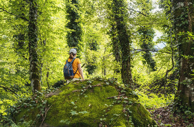 Rear view of woman with backpack walking in summer forest next to green moss stone in summer