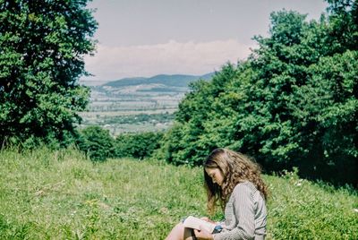Young woman reading book while sitting on grassy field against trees