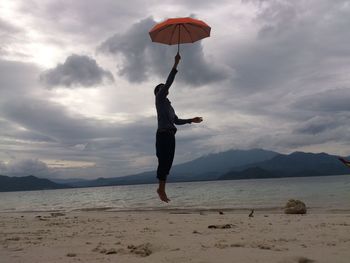 Side view of young man with umbrella jumping at beach against cloudy sky
