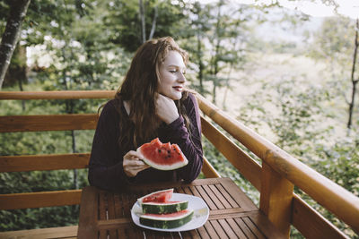 Young redhead woman eating watermelon on wooden house balcony