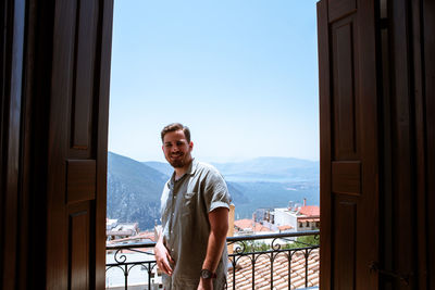 Young male standing on wooden balcony near the mountains and smiling at the camera.