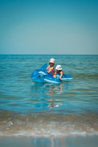 Siblings on inflatable rafts in sea against blue sky