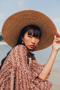 Portrait of young woman wearing hat sitting at beach against sky