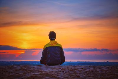 Rear view of man sitting at beach against sky during sunset