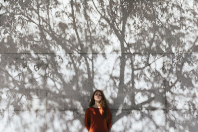 Young woman with hands behind back standing against tree shadow wall