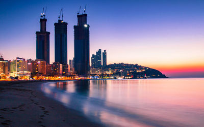 Illuminated buildings by sea against sky at dusk